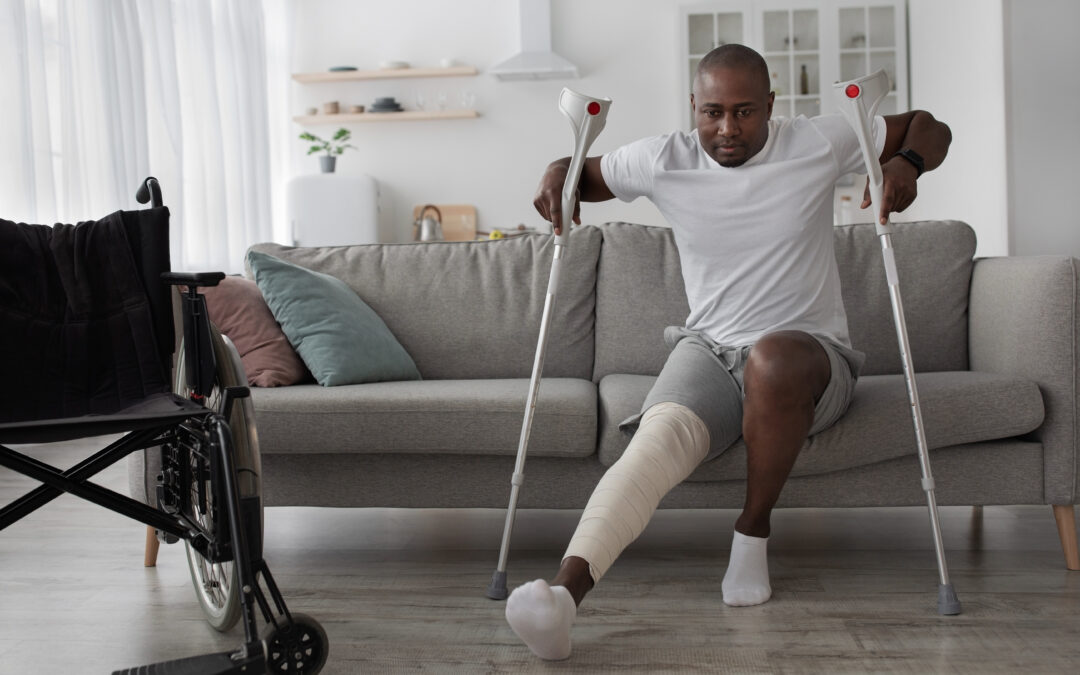 Black middle aged male with broken leg with plaster cast, recovering from orthopedic injury, gets up from sofa with crutches in living room interior with wheelchair.